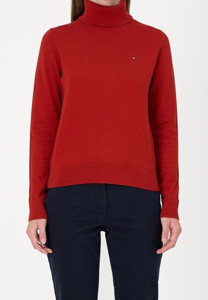 Mujer con un suéter ajustado de cuello alto rojo de manga larga y pantalones azul marino, de pie frente a un fondo liso.