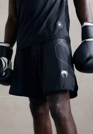 Boxer wearing black gloves, black shorts with a white snake logo, and a black shirt, standing against a plain background.