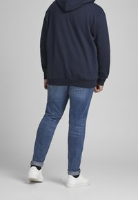 Felpa con cappuccio di colore navy, con polsini e orlo a coste, abbinata a jeans in denim blu chiaro. Sneakers bianche visibili ai piedi.