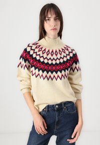 Knit sweater with a high neck, cream body, and colorful geometric pattern in red, navy, and pink. Ribbed cuffs and hem. Worn with dark jeans.
