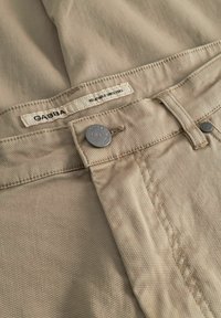 Bézs színű, texturált szövetszövetből készült nadrág fém gombbal és övbújtatóval, melyen "GABBA" és "RELATABLE SINCE 1983" felirat található.