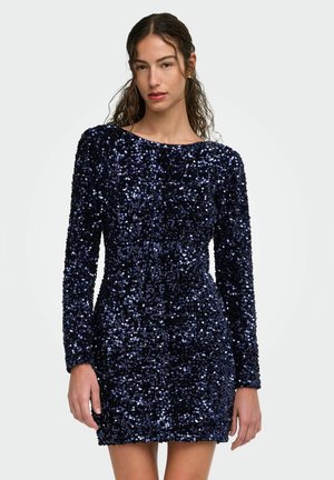 Femme portant une mini-robe à manches longues en sequins bleu marine avec un col rond, debout devant un fond clair uni.