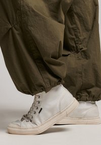 Witte canvas hoge sneakers met een gestructureerde rubberen zool, gecombineerd met losse olijfgroene joggingbroeken die bij de enkels zijn samengebonden.