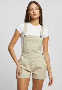 Beige denim overaller med justerbare stropper og en frontlomme. Har sølvfarget utstyr og oppbrettede mansjetter ved benåpningene.