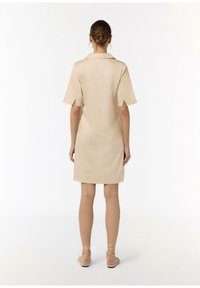 Vestido de camisa beige con mangas cortas, cuello y lazos en los extremos de las mangas. Textura suave, hasta la rodilla, combinado con sandalias de color claro.
