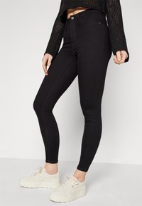 Mujer vestida con jeans skinny de tiro alto negros y zapatillas Puma blancas, de pie contra un fondo claro y liso.