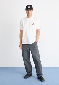 Uomo in piedi su un pavimento blu che indossa una maglietta bianca con un piccolo logo nero, pantaloni grigi, sneakers nere e un cappellino nero.