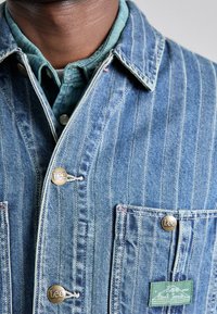 Jeansjakke med lodrette hvide striber, to frontlommer og bronzefarvede knapper. Har et grønt mærkat på venstre lomme.