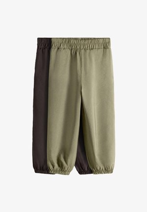 Fuß in braunem Wildleder-Mary-Jane-Schuh mit weißem Besatz, kombiniert mit locker sitzenden olivgrünen Hosen, die am Knöchel gerafft sind, stehend auf hellem Boden.