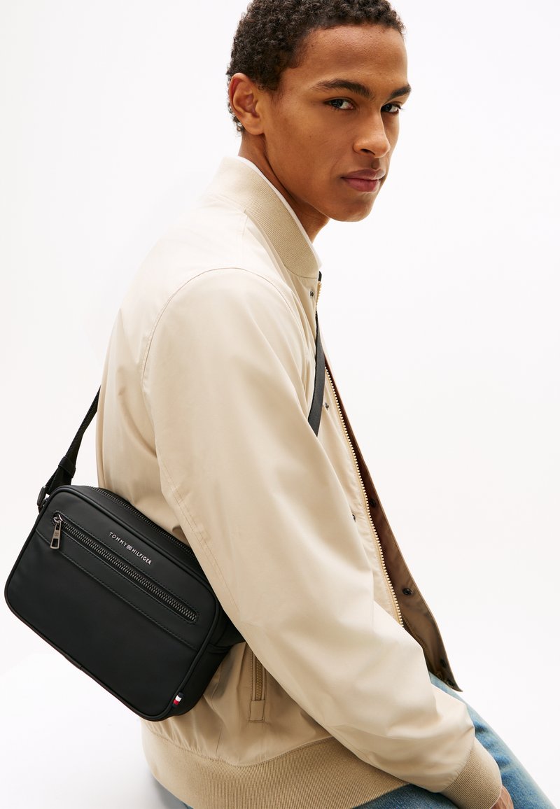 Νέος άνδρας με μπεζ μπουφάν και τζιν φορεί μια μαύρη τσάντα Tommy Hilfiger crossbody σε λευκό φόντο.