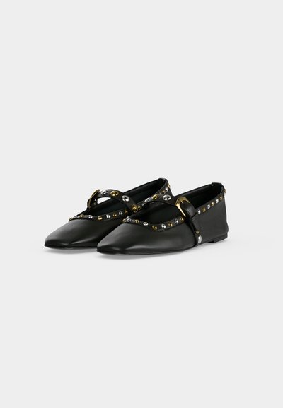 Paire de ballerines noires à bout pointu, avec brides ornées de clous dorés et argentés, et une boucle dorée, présentées sur un fond uni clair.