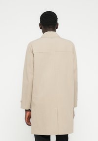 Μπεζ, μακρύ παλτό με ευθεία γραμμή, ελάχιστες ραφές και πίσω καβάλο. Διαθέτει μακριά μανίκια και κουμπιά στα μανίκια.