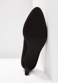Chaussure femme noire à bout pointu avec semelle texturée, petit talon et logo Clarks visible au-dessous.
