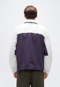 Άνδρας που φοράει λευκό και σκούρο μωβ μπουφάν και σκούρο παντελόνι, στέκεται γυρισμένος με την πλάτη σε λευκό, απλό φόντο.