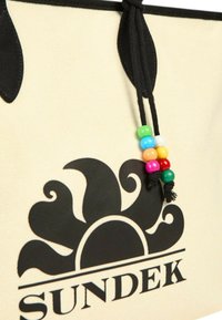 Borsa tote in tela color crema con logo nero e design a onde. Presenta manici neri e un accento decorativo di perline colorate.
