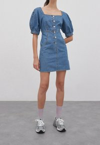 Denim mini-mekko, jossa on puffatut lyhyet hihat, neliön muotoinen kaula-aukko, etusuljenta nappikiinnityksellä ja näkyvillä ompeleilla. Yhdistetty harmaisiin lenkkikenkiin ja violetteihin sukkiin.