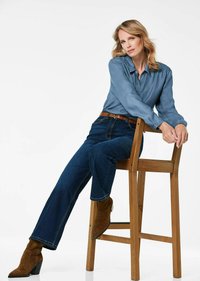 Helles blaues Denimhemd mit lockerer Passform, kombiniert mit dunkelblauen Schlagjeans und braunen Wildleder-Ankle-Boots. Holzstuhl im Hintergrund.