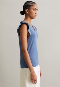 Blaues ärmelloses Top mit Rüschendetails an den Schultern, V-Ausschnitt und glattem Baumwollstoff, kombiniert mit hellen Hosen.