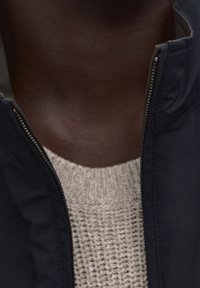 Navy zippered jacket made of smooth fabric, layered over a light beige knitted sweater with a textured pattern.