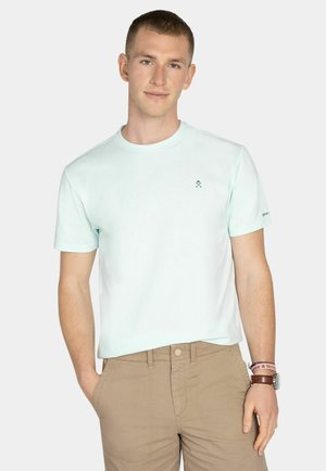 Joven con camiseta de manga corta azul claro y pantalones caqui, de pie con una mano en el bolsillo frente a un fondo liso.