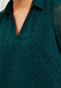 Teal blouse with a collar, featuring a subtle floral pattern in dark blue. Soft fabric with a relaxed fit and a partially open neckline.