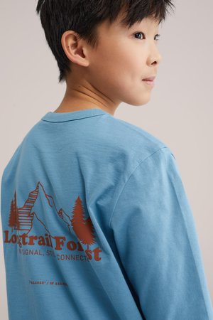 Mladi dječak u plavoj majici s grafikom planine i stabla na leđima s natpisom "Longtrail Forest, nema signala, ali još uvijek povezan", gleda prema desno.