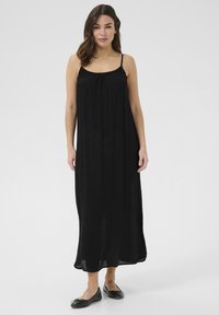 Vestido maxi negro de tejido ligero con tirantes finos y pecho fruncido. Zapatos planos negros complementan el atuendo, ofreciendo un aspecto casual.
