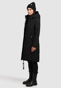 Abrigo acolchado largo negro con capucha ajustada, diseño acolchado, bolsillos laterales y tirador de cremallera con marca, combinado con botas negras de tobillo y un gorro.