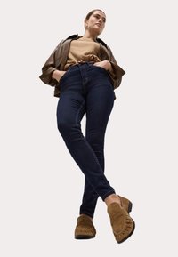 Jeans de talle alto de color azul oscuro, chaqueta de cuero marrón, suéter color topo y zapatos de estilo mocasín marrones con detalles de flecos.