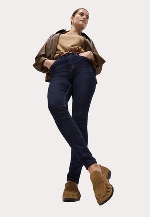Jeans taille haute bleu foncé, veste en cuir marron, pull beige, et chaussures mocassins marron avec des détails frangés.