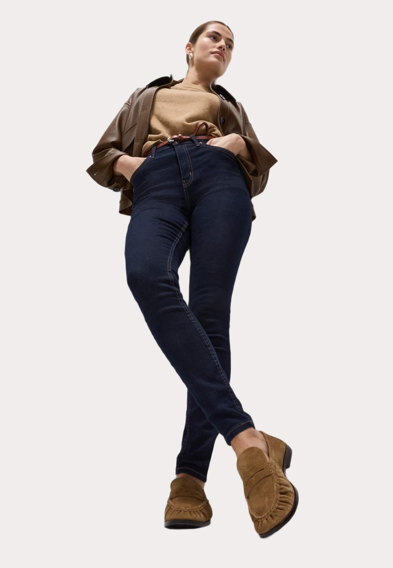 Jeans de talle alto de color azul oscuro, chaqueta de cuero marrón, suéter color topo y zapatos de estilo mocasín marrones con detalles de flecos.