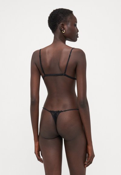 Conjunto de lencería de encaje negro que presenta tirantes finos, un diseño transparente y acentos florales en el tanga. Mostrado desde un ángulo trasero.