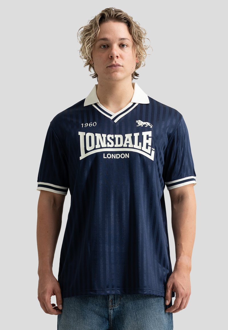 Jeune homme aux cheveux blonds bouclés portant un maillot de sport bleu marine Lonsdale London avec col blanc et bandes.