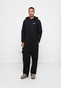 Sudadera negra con bolsillo frontal y detalle de logo, combinada con pantalones cargo negros de corte suelto y botas grises. El fondo es blanco liso.