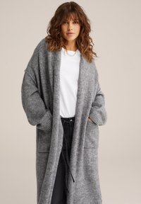 Pelēks garš kardigans, kas izgatavots no mīkstas, strukturētas adītas auduma ar brīvu piegriezumu, platām piedurknēm un divām priekšējām kabatām. Apvienots ar baltu kreklu.