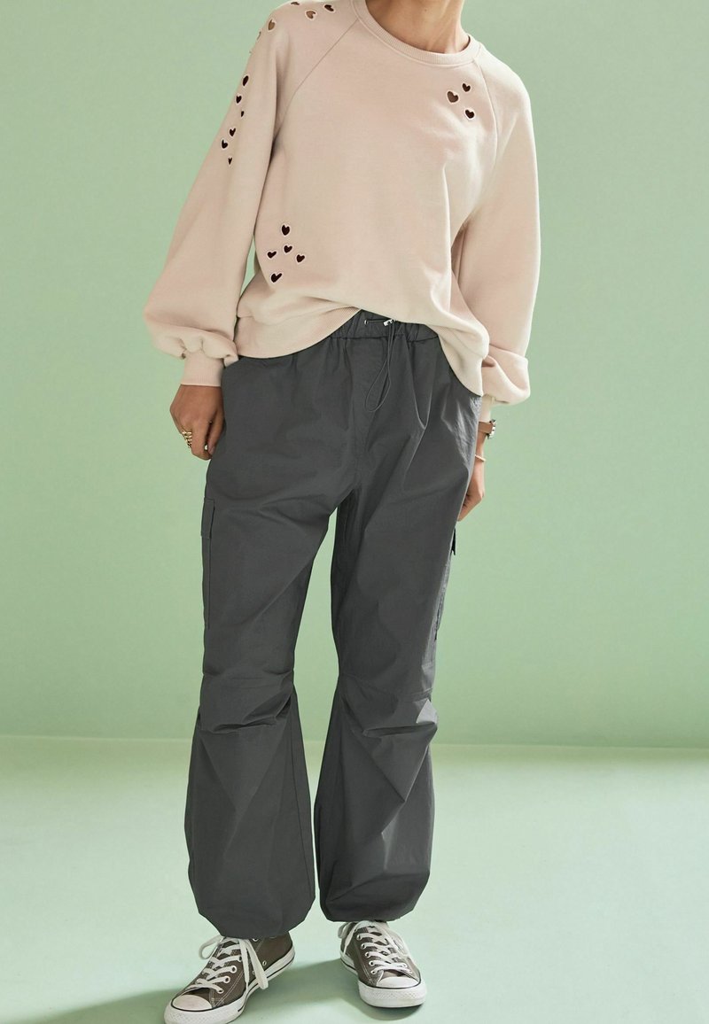 Felpa rosa chiaro con tagli a forma di cuore sulle maniche, abbinata a pantaloni cargo grigi e larghi e sneakers marroni. Design semplice, stile casual.