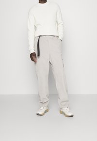 Hellgraue Cargohosen mit schwarzem Gürtel und seitlichen Taschen, kombiniert mit einem weißen, strukturierten Pullover und mehrfarbigen Sneakern.