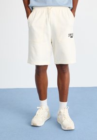 Shorts couleur crème en tissu doux, dotés de poches latérales et d'un logo visible. Associés à des chaussures et des chaussettes de couleur claire.