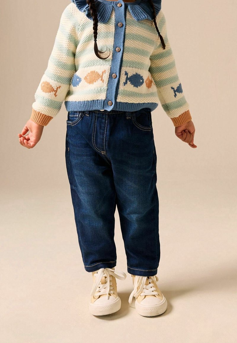 Enfant portant un pull rayé avec des motifs de poissons, un jean bleu foncé et des baskets crème, debout devant un fond uni.