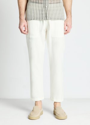 Uomo che indossa pantaloni bianchi a gamba dritta con tasche frontali e scarpe beige slip-on, in piedi su uno sfondo chiaro e uniforme.