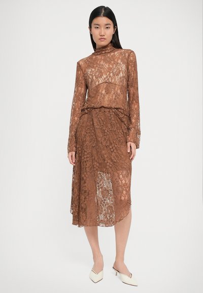Robe en dentelle marron avec des manches longues et un col haut. Présente des motifs floraux et un design superposé, assortie à des chaussures de couleur crème.