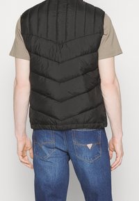 Person, der eine schwarze gepolsterte Weste über einem beigen kurzärmligen Hemd und blauen Jeans trägt, die sichtbare Gesäßtaschen und ein Markenlogo aufweisen.