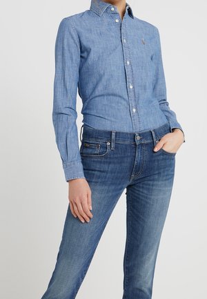 Kvinde iført en tætsiddende lyseblå skjorte med knapper, stoppet ned i mellemhøje mørkeblå jeans, med den ene hånd i lommen mod en ensfarvet baggrund.