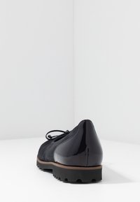 Czarne damskie płaskie buty z zamszowym przodem, lakierowanym obcasem, brązowymi wykończeniami i podeszwą z rowkami, fotografowane od tyłu na białym tle.