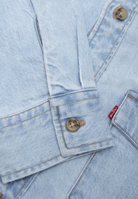 Light blue denim jacket featuring dual buttons, contrasting stitching, and a red brand tag, with a structured collar and cuff detail.