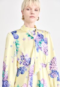 Geel bloemenjurk met paarse en blauwe hortensia-print, satijnen stof, lange mouwen en een strikdetail bij de hals. Textuurmet bloemenaccenten zichtbaar.