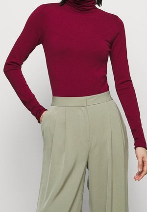 Personne portant un haut à col roulé bordeaux ajusté, rentré dans un pantalon large plissé gris clair à taille haute, une main dans la poche.