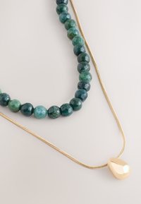 Zwei Halsketten: eine mit dunkelgrünen, facettierten Perlen, die andere eine dünne Goldkette mit einem glatten, tropfenförmigen Anhänger.