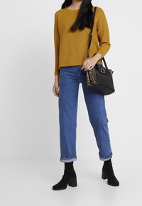 Gelber gestrickter Pullover mit langen Ärmeln, kombiniert mit mittel-hoch geschnittenen blauen Jeans. Schwarze Absatzstiefel und eine schwarze Tasche mit einem gemusterten Seitenteil.