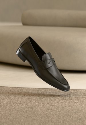 Zapato mocasín de cuero negro flotando sobre una alfombra beige con un sofá beige en un ambiente interior minimalista.